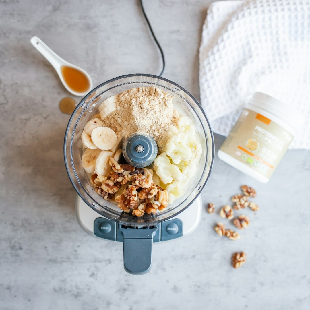 Bananaer og nødder der ligger i en blender og en bøtte med protein ved siden af.